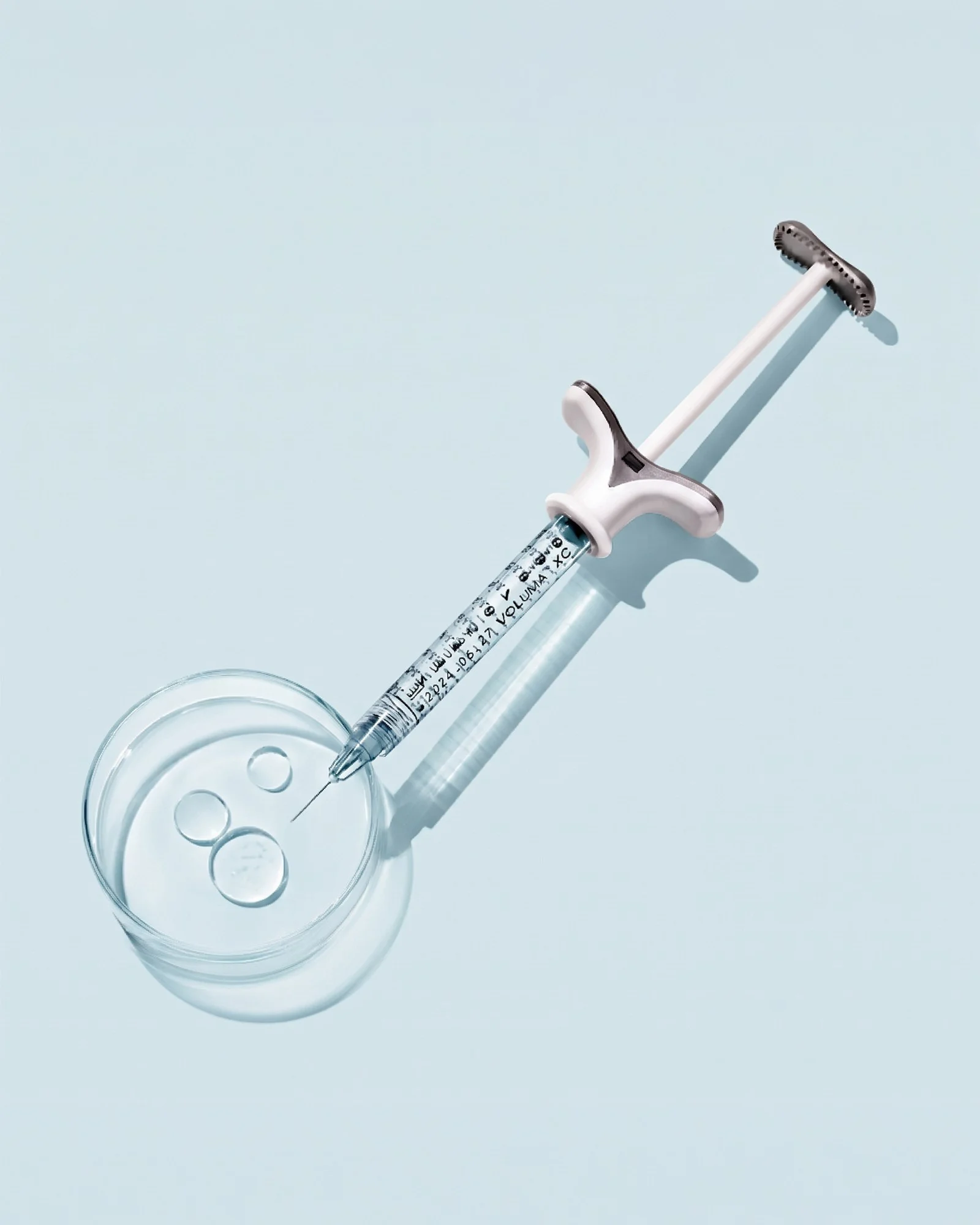 A syringe of Juvéderm Voluma XC filler beside a dish of clear gel droplets
